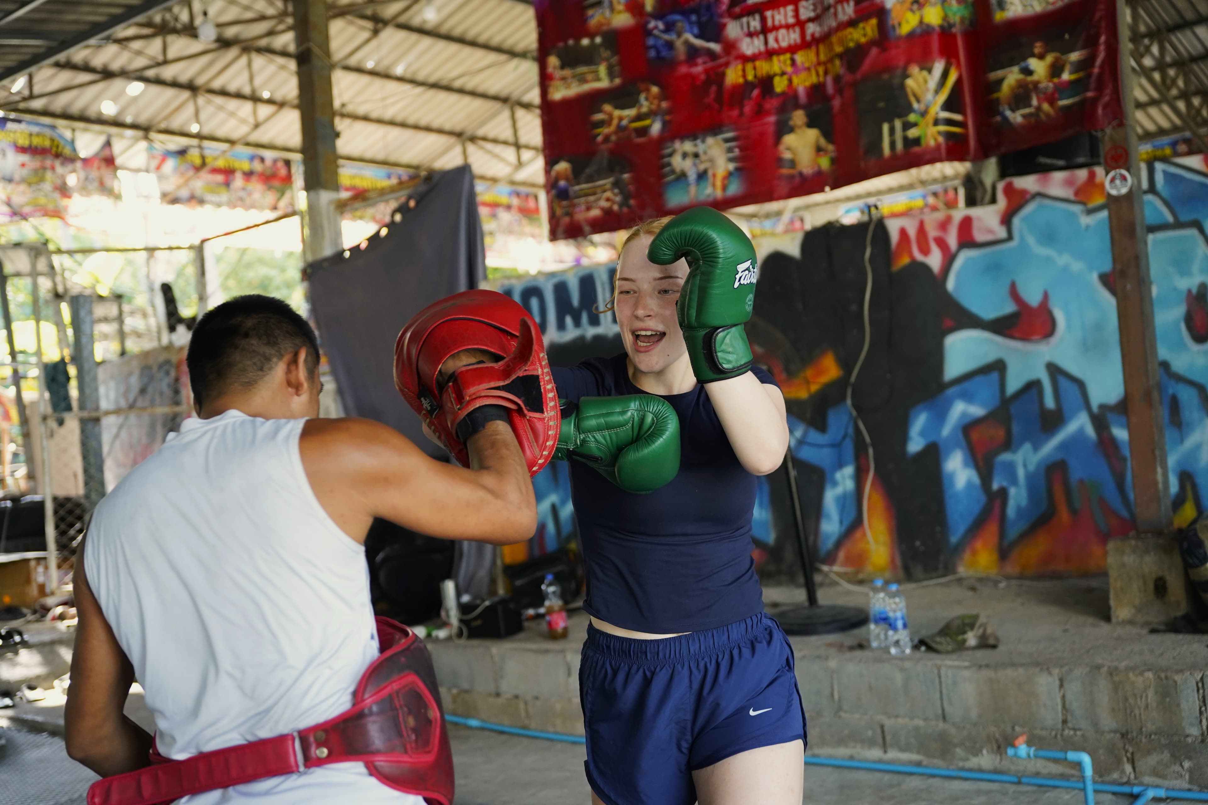 A trainer holding focus mitts for a woman throwing a punch at an outdoor gym with a graffiti wall