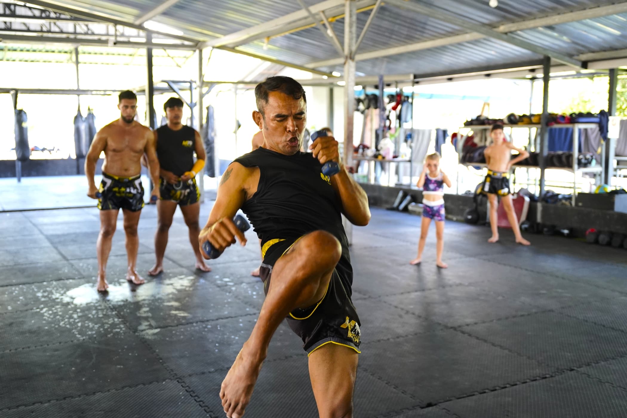 A Thai trainer demonstrating a knee strike while a group of students watches at a gym